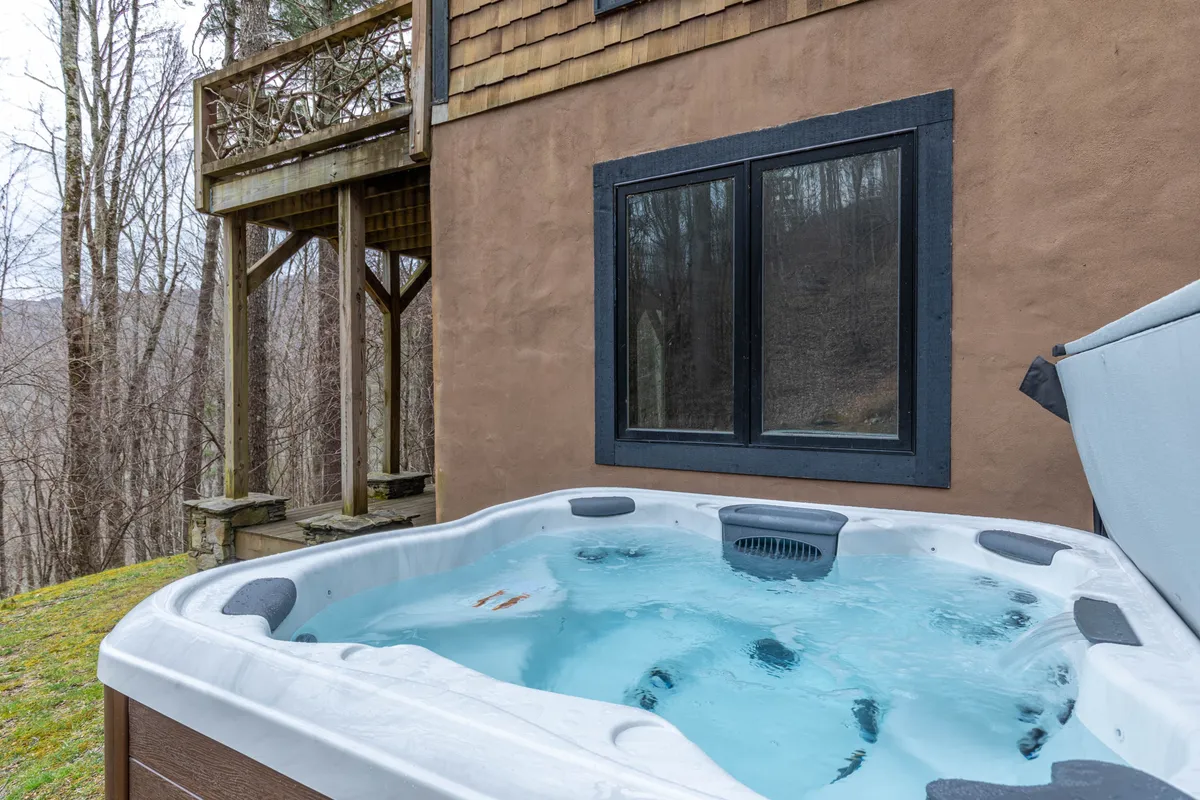 This exterior image shows a private hot tub set on a wooden deck, surrounded by trees, with a window offering a glimpse into the home. The setting is tranquil and private, ideal for relaxing soaks under the stars or after a long day outdoors.