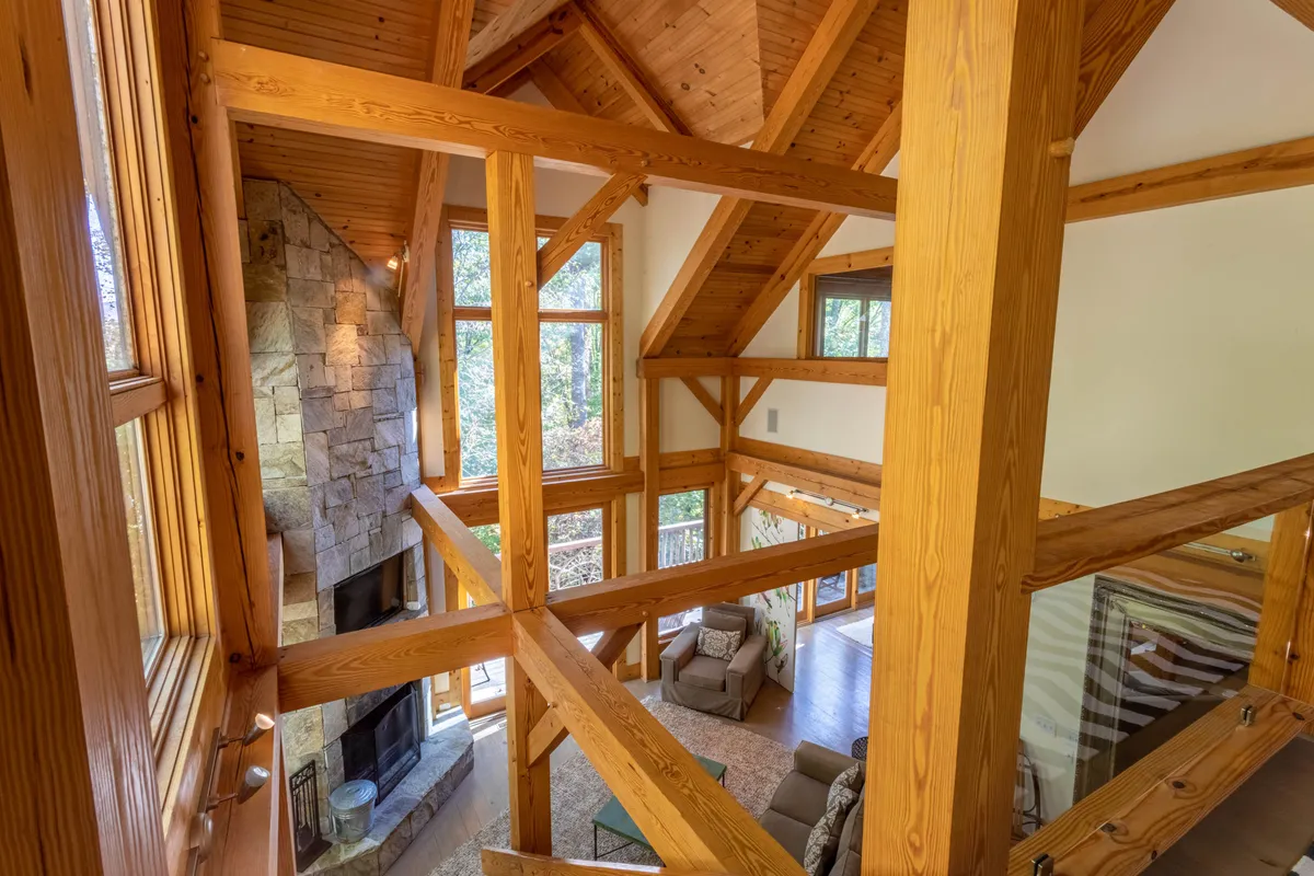 An interior view featuring an expansive living area with a large stone fireplace, exposed wooden beams, and dramatic floor-to-ceiling windows offering breathtaking views of the natural surroundings. The furnishings encourage cozy gatherings and relaxation by the fire while enjoying the mountain scenery.