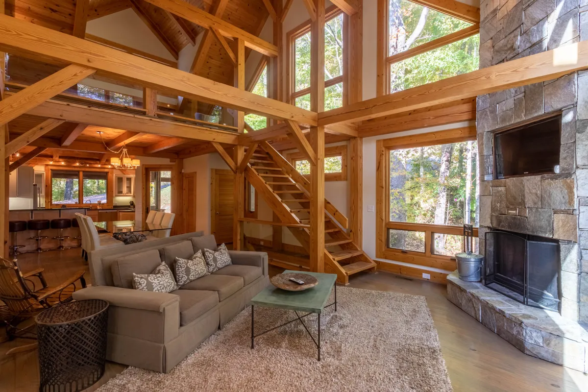 A spacious Great Room featuring expansive windows with sweeping mountain views, rustic timber beams overhead, a dramatic floor-to-ceiling stone fireplace as the focal point, and a plush, comfortable sofa inviting relaxation and conversation. Soft daylight streams in, creating a warm, inviting ambiance perfect for gatherings.