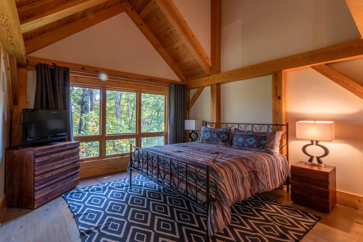 A cozy bedroom with a plush bed, decorative throw pillows, large windows offering views of nature, a stylish bedside lamp, and a wooden dresser. The room exudes warmth and privacy, perfect for couples or friends seeking a private retreat.
