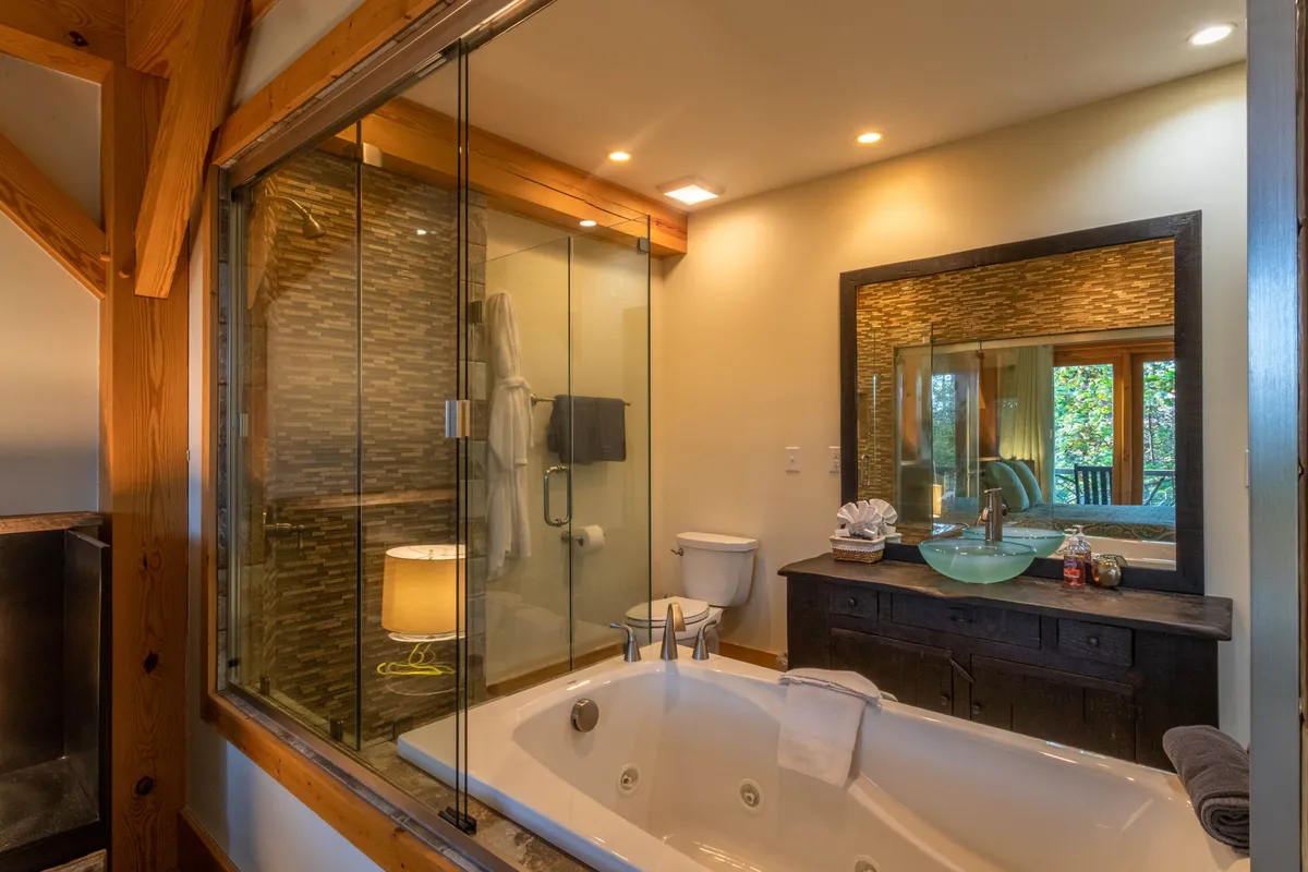 A luxurious ensuite bathroom with a jacuzzi bathtub, glass shower enclosure, large mirror, and warm wooden vanity. The decor is calming and spa-like, echoing mountain lodge inspiration with its use of wood and earth tones.