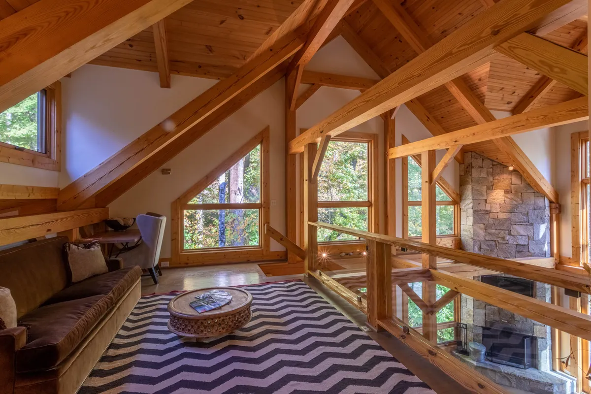 This image shows a spacious living area featuring large windows, exposed wooden beams, a stone fireplace, a zigzag patterned rug, and a comfortable sofa. The windows provide ample views of the outdoors, connecting the cozy indoor environment to the natural landscape beyond.