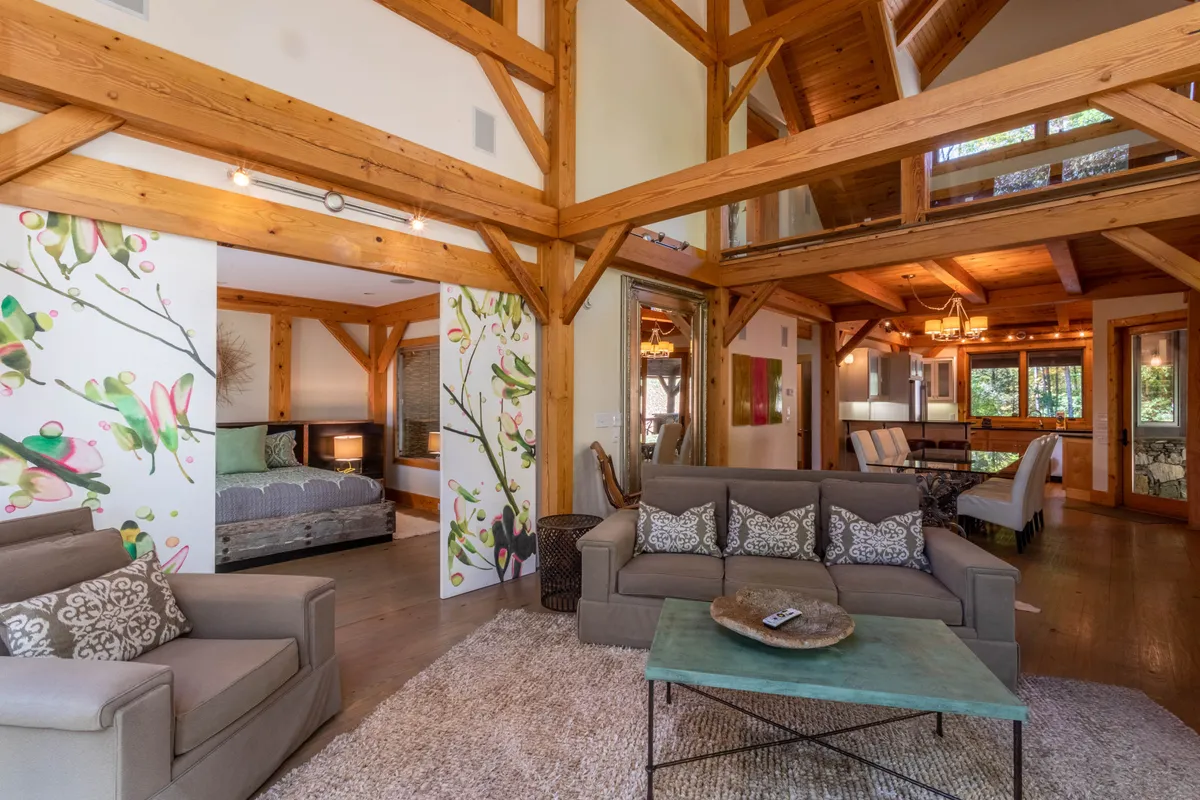 This image highlights a cozy living space with wooden beams, a sofa adorned with patterned cushions, and colorful wall art. Light streams in, creating a sunny, inviting atmosphere ideal for morning coffee or unwinding with a pet.