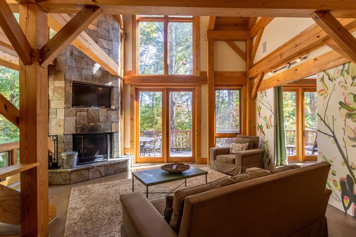 The image showcases a stunning living area featuring a towering stone fireplace, expansive floor-to-ceiling windows framing mountain views, rustic wooden beams, and plush seating arranged for comfort. Sunlight pours in, illuminating the cozy yet airy space, and inviting guests to relax while taking in the natural beauty outside.