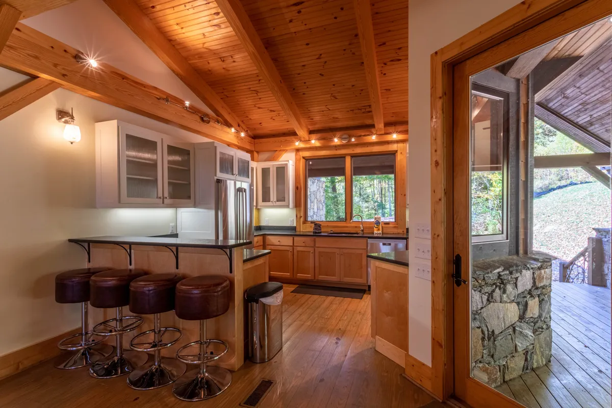An open, inviting kitchen with a large island and bar stools, surrounded by wooden beams and stone accents. Ample windows provide a backdrop of natural light, making this space ideal for cooking, entertaining, and sharing meals with family and friends.