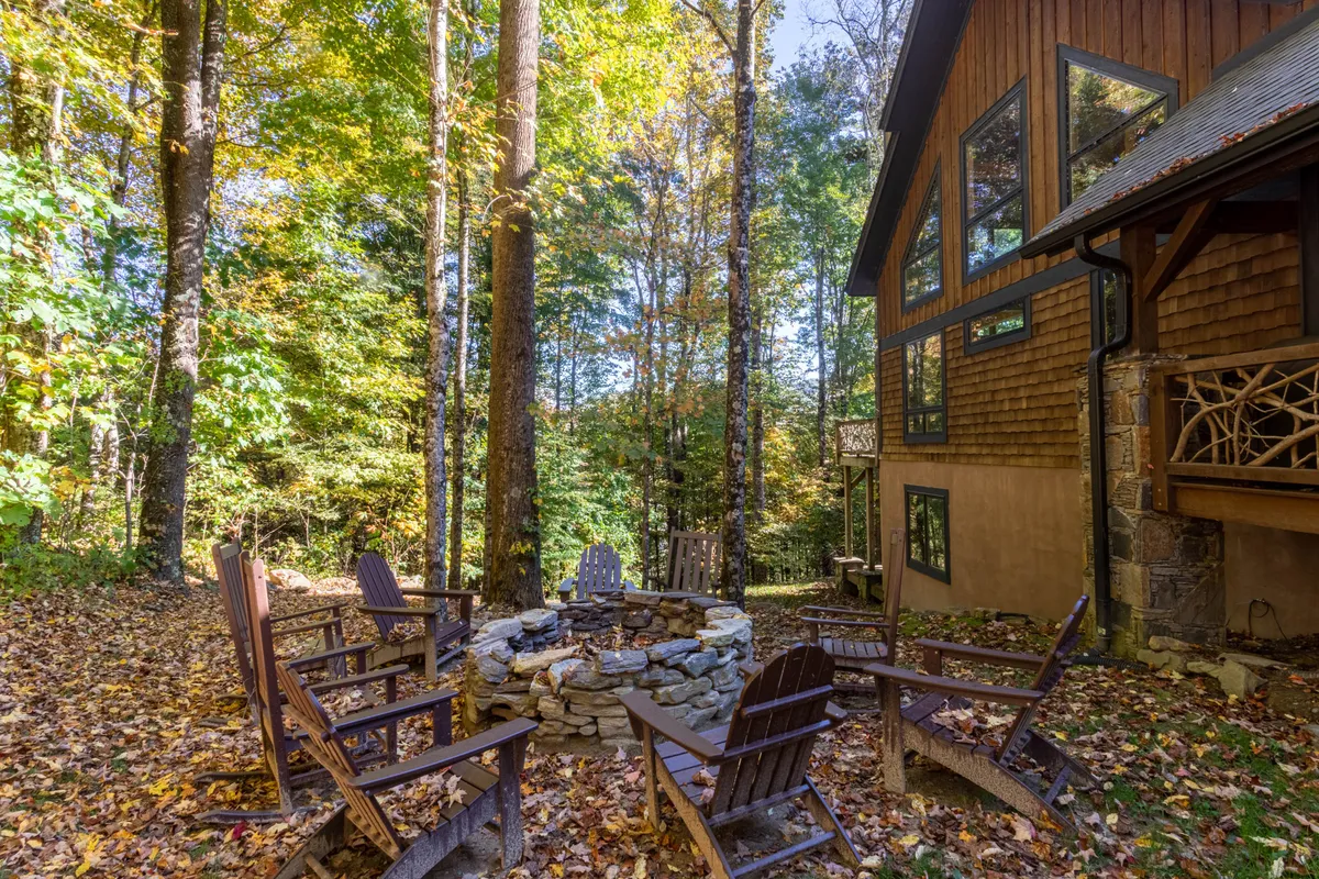 A cozy outdoor fire pit surrounded by comfortable chairs, set amid tall trees with a view of the house exterior. The setting radiates warmth and invites group gatherings, with the forested environment enhancing the feeling of a mountain retreat.