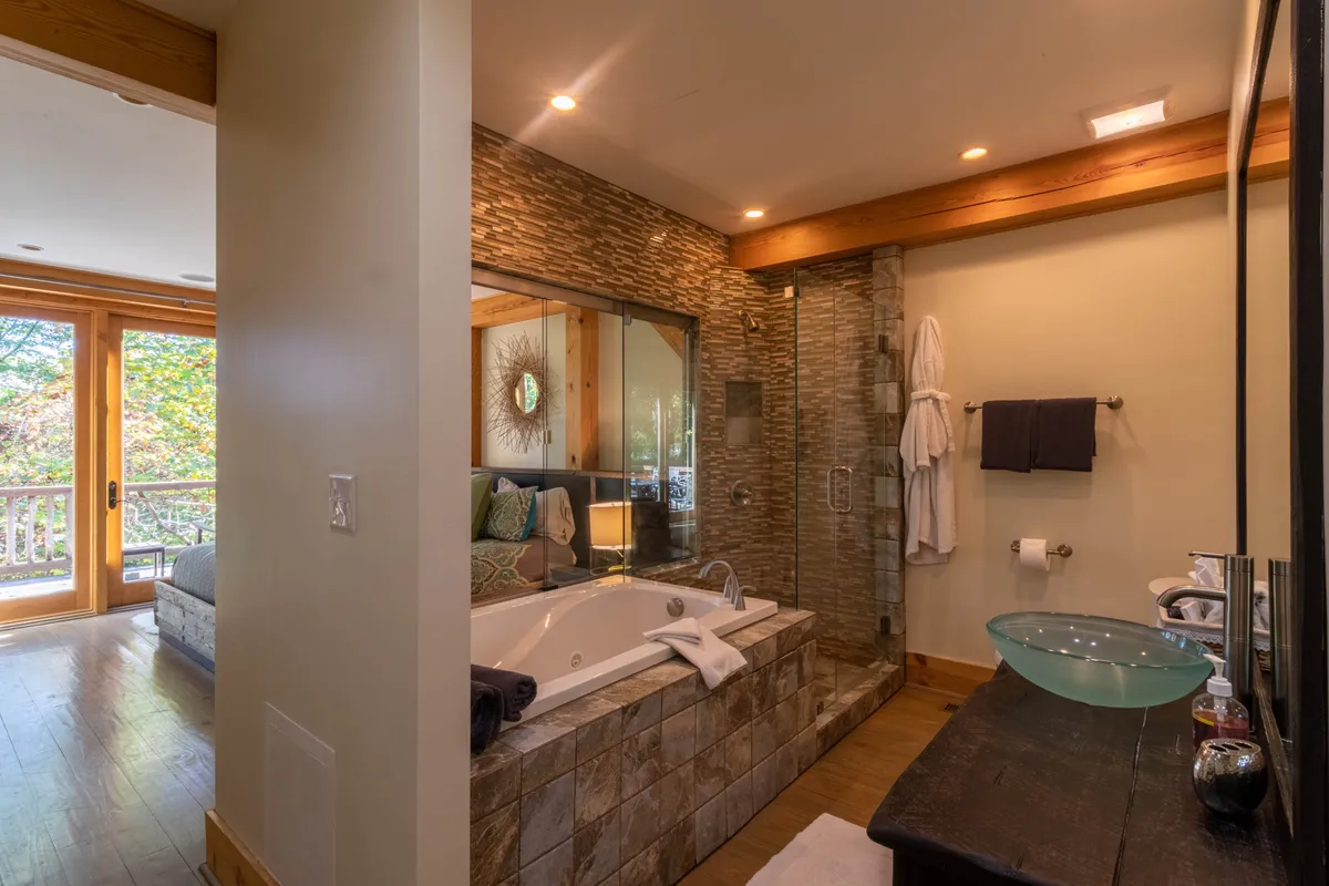 A luxurious bathroom with a jacuzzi tub set beside large glass windows, wooden accents, and a decorative mirror. The tub overlooks natural scenery, and the space exudes a spa-like atmosphere—perfect for relaxing after a day of mountain activities.