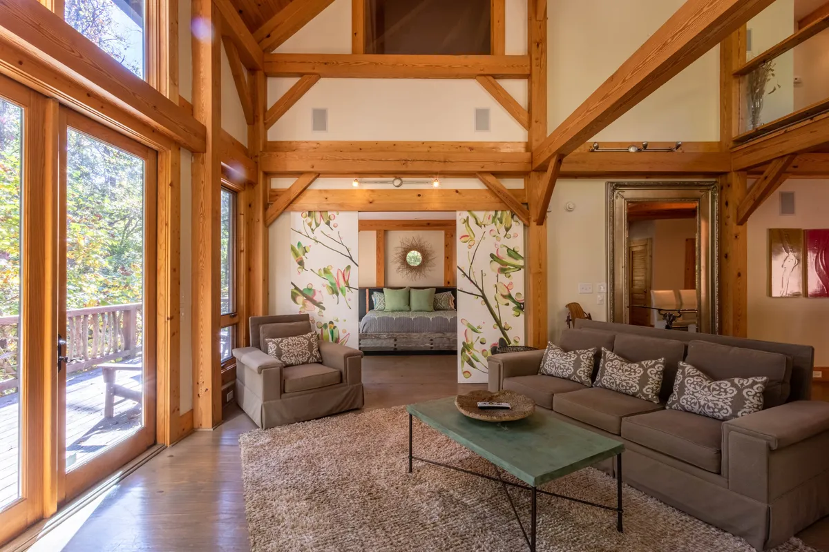 This scene captures a welcoming seating area beneath exposed wooden beams, with floral wall art and large glass doors that likely open onto a patio or deck. The space is filled with natural light, offering a glimpse of the outdoors.