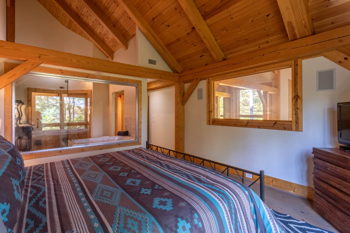 A cozy bedroom with a bed featuring a patterned blanket sits beside a large window. Natural light floods the room, and exposed wooden beams add rustic charm. The window hints at an inviting view of the outdoors, perfect for stargazing or early morning light.