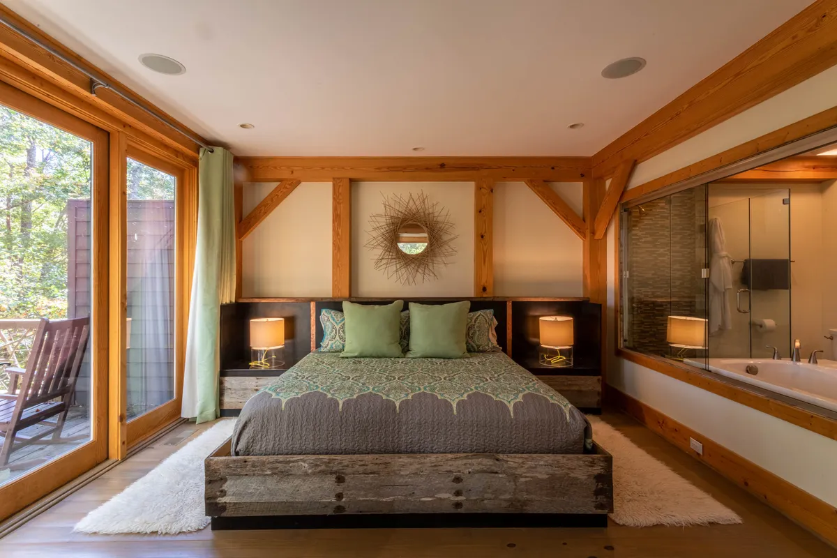 A serene master suite featuring a comfortable bed positioned near large windows that overlook nature. The room includes a decorative mirror and an ensuite bathroom area visible in the background. Soft natural light fills the space, and mountain-inspired finishes and calming tones create an atmosphere of relaxation.