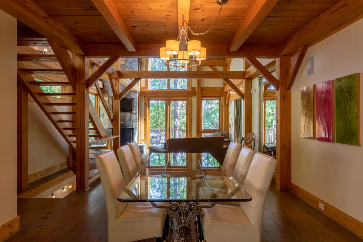 A welcoming dining area featuring a beautiful wooden table under an elegant chandelier, timber beams overhead, and large windows offering glimpses of the outdoors. The setting is bright and airy, ideal for festive celebrations or intimate family dinners.